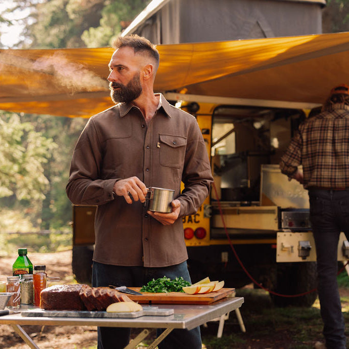 A bearded man in a brown wool button-up heritage shirt stands outdoors under a yellow canopy next to a camper, holding a metal pot. A folding table in front holds food items, and another person is visible working by the camper in a wooded setting.