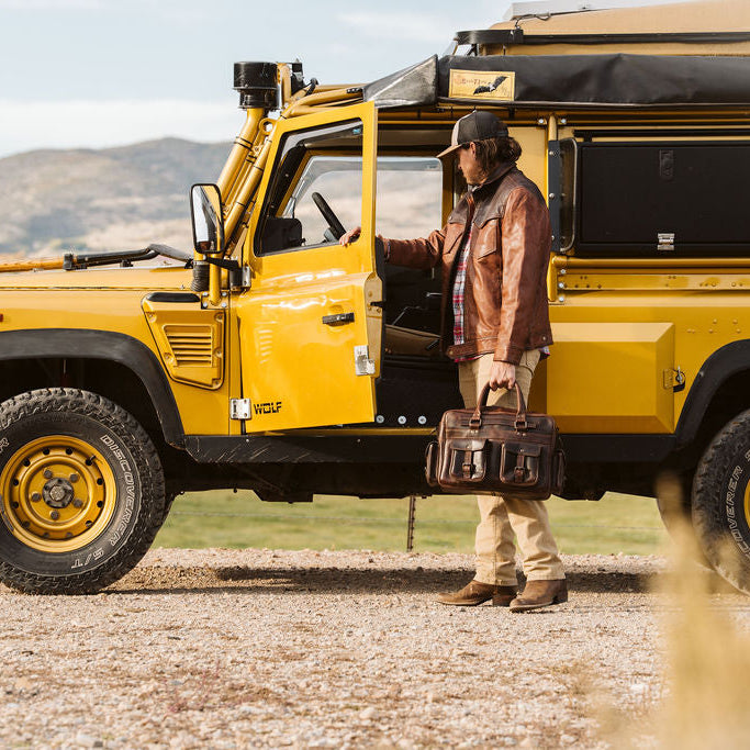 A man in a brown leather jacket and beige pants stands beside a bright yellow off-road safari vehicle, Land Rover Wolf, holding a brown leather briefcase in an outdoor desert setting.