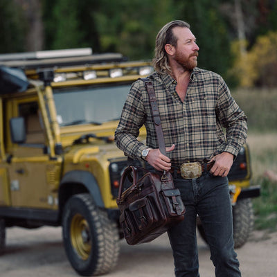 A man wearing a brown and beige plaid shirt and dark jeans stands on a dirt road next to a yellow off-road vehicle, carrying a dark brown leather shoulder bag.