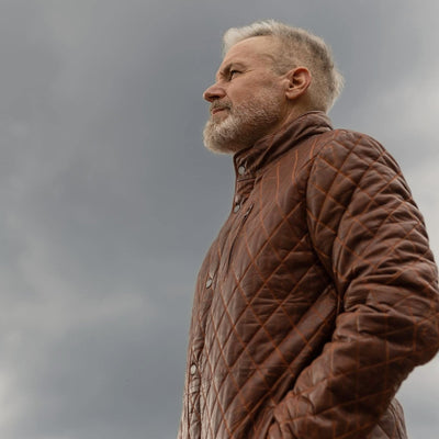 Man in the Highlands Quilted Leather Jacket in Whiskey Brown, side view under a stormy sky.