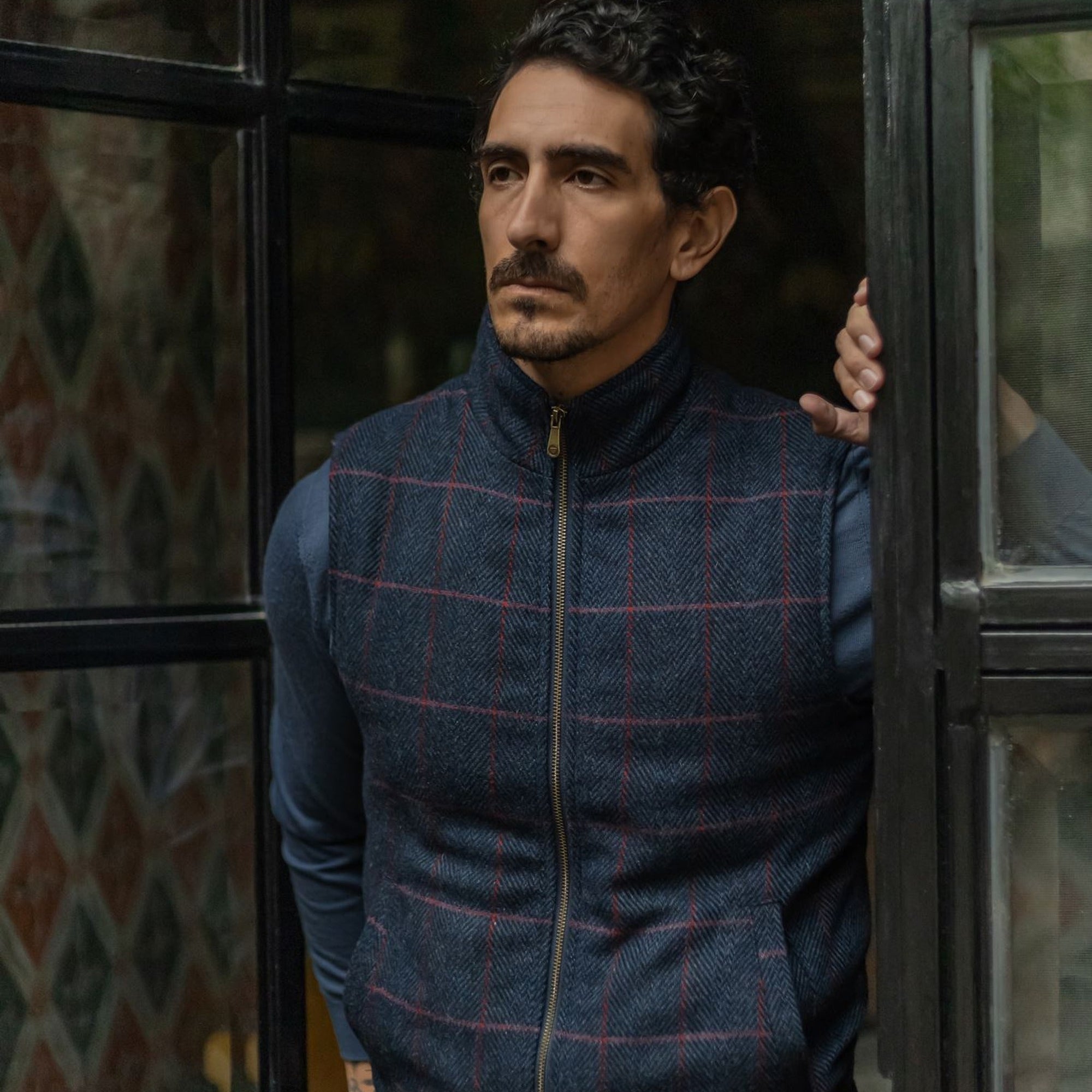 Man in the Ashton Wool Herringbone Vest in Clan Blue standing at a dark steel window, hand on the frame