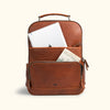 Brown leather backpack with front zipper pocket and open compartment storing a white tablet and travel postcard.