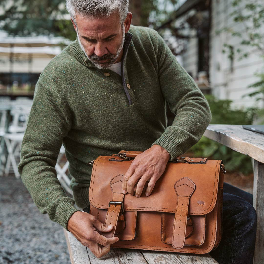 Mature man in green sweater using Roosevelt amber brown leather briefcase on a rustic wooden table.