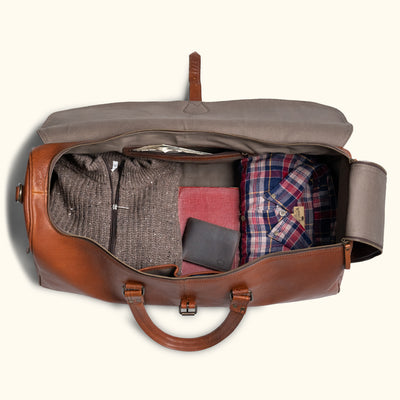 Open vintage brown leather duffle bag packed with clothes, showing a plaid shirt, sweater, and wallet in organized compartments.