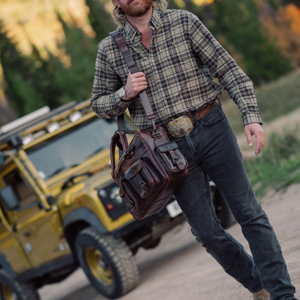 A man with a beard wearing a green and brown plaid shirt, dark jeans, and boots walks along a dirt road carrying a brown leather messenger bag over his shoulder. A yellow off-road vehicle is parked in the background against a blurred forest backdrop.