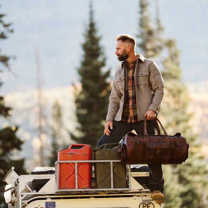 A man stands on the back of a vehicle in a forested outdoor setting, holding a brown duffel bag. He is wearing a brown overshirt over a plaid shirt and dark pants, with a red fuel canister and other bags strapped to the vehicle rack.