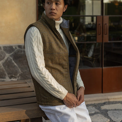 Man in the Ashton Wool Herringbone Vest in Mosswood seated on a wooden bench outside a cafe
