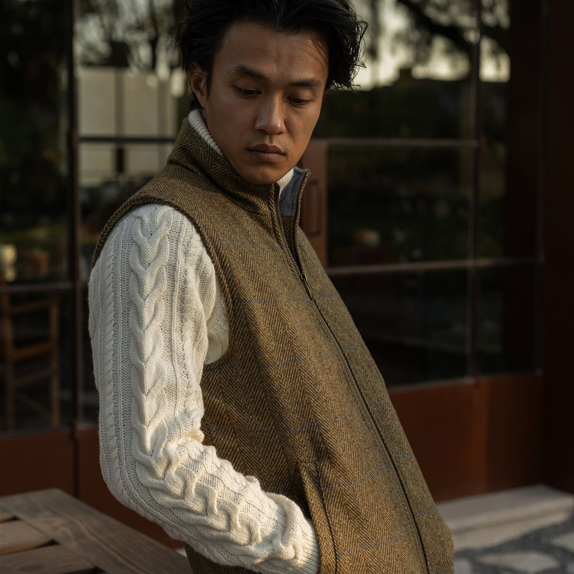 Man in the Ashton Wool Herringbone Vest in Mosswood over a cream cable-knit sweater on a terrace