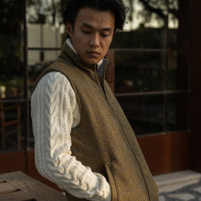 Man in the Ashton Wool Herringbone Vest in Mosswood over a cream cable-knit sweater on a terrace