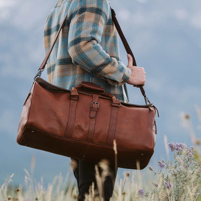 Rustic brown leather travel bag showcased by an adventurer in the natural beauty of Jackson Hole, Wyoming, perfect for outdoor expeditions