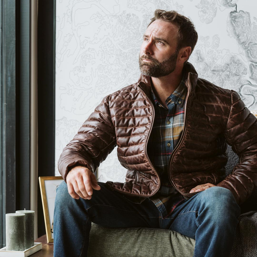 Casual look: man in a brown quilted jacket and jeans, sitting indoors with a contemplative expression.