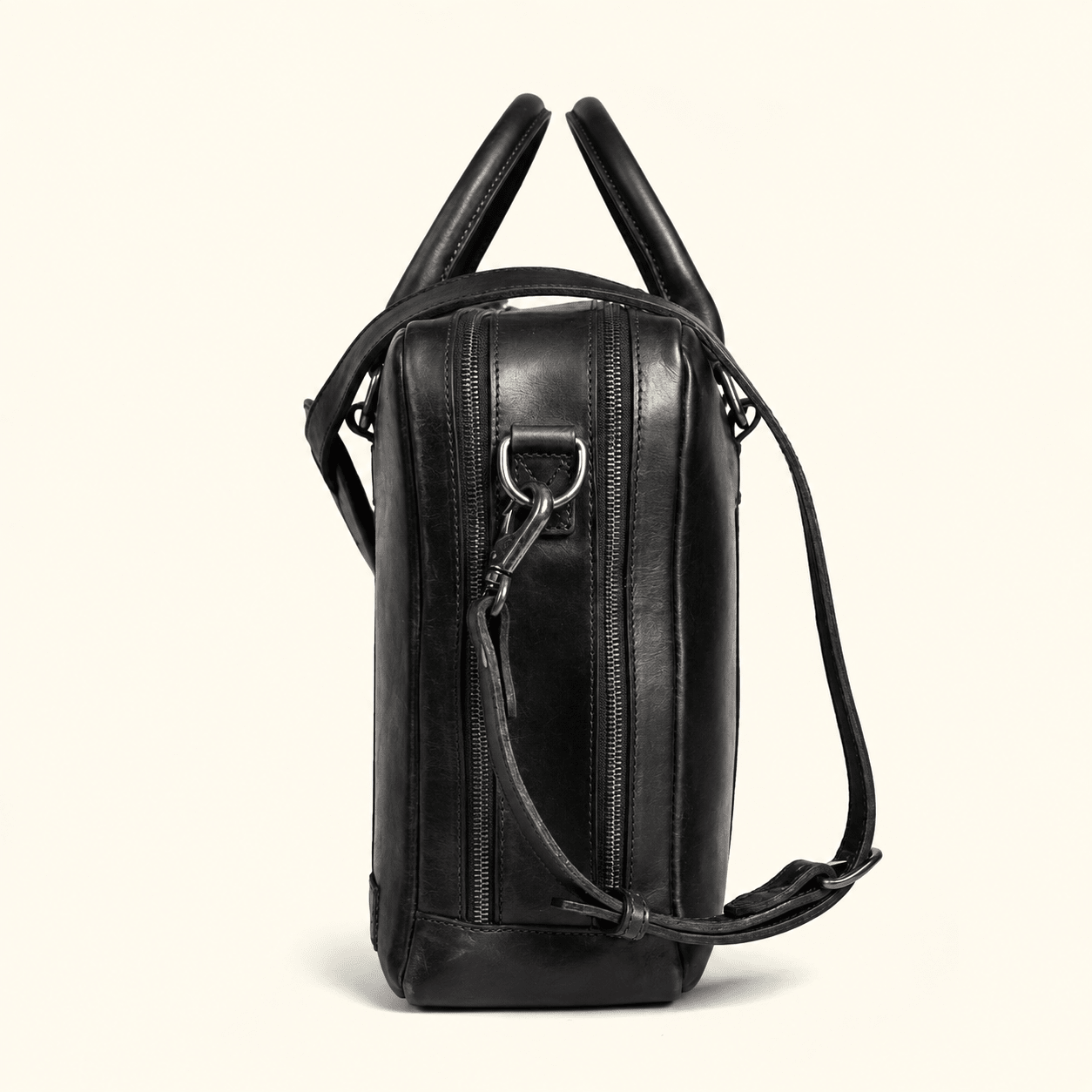 A black leather shoulder bag with dual top handles and a detachable shoulder strap, shown from a side profile view against a light cream background.