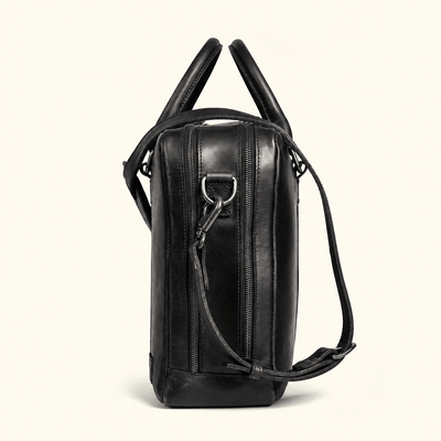 A black leather shoulder bag with dual top handles and a detachable shoulder strap, shown from a side profile view against a light cream background.