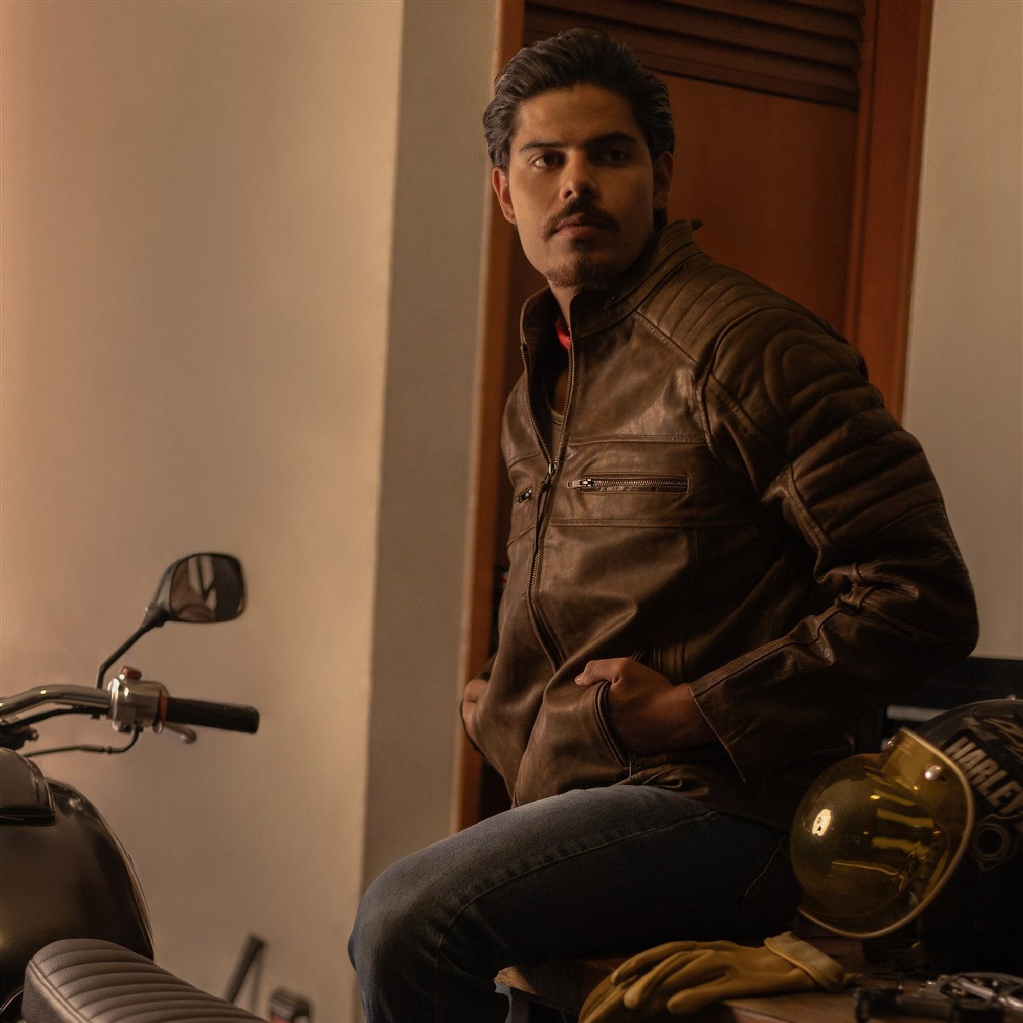 Man in the Cafe Racer Leather Jacket in Distressed Brown seated by his motorcycle in a garage, jacket front in view.