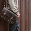 Man holding a dark oak Roosevelt leather briefcase with antique brass buckles and adjustable shoulder strap.