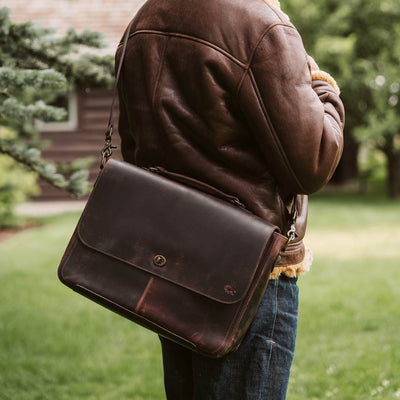 Man wearing a leather jacket and jeans carries a distressed dark brown leather messenger bag with a secure back pocket zipper in a lush garden setting.
