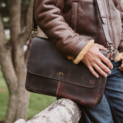 Man outdoors wearing a leather jacket, casually holding a dark brown distressed leather messenger bag with visible wear and a sturdy shoulder strap.