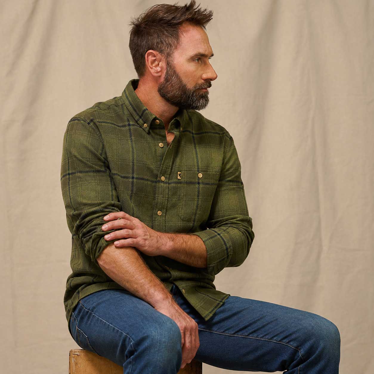A man wearing an olive green plaid button-down shirt and blue jeans, sitting on a wooden stool against a neutral beige fabric backdrop.