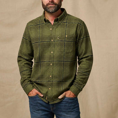 A man wearing an olive green plaid button-down shirt and blue jeans, standing against a beige fabric backdrop.