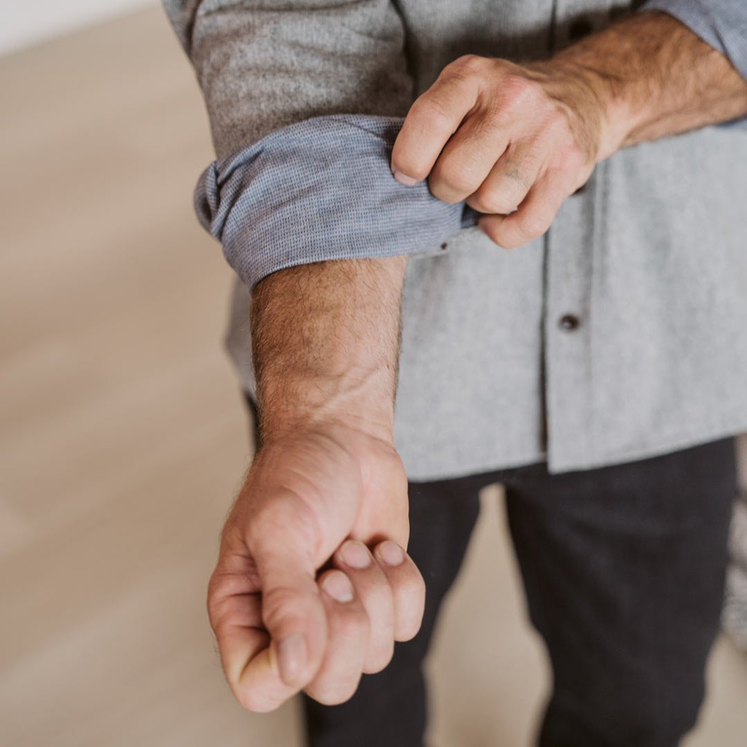 A close-up of a man's hands rolling up the sleeve of a heather gray long-sleeve button-up shirt, paired with dark trousers, in a brightly lit indoor setting.