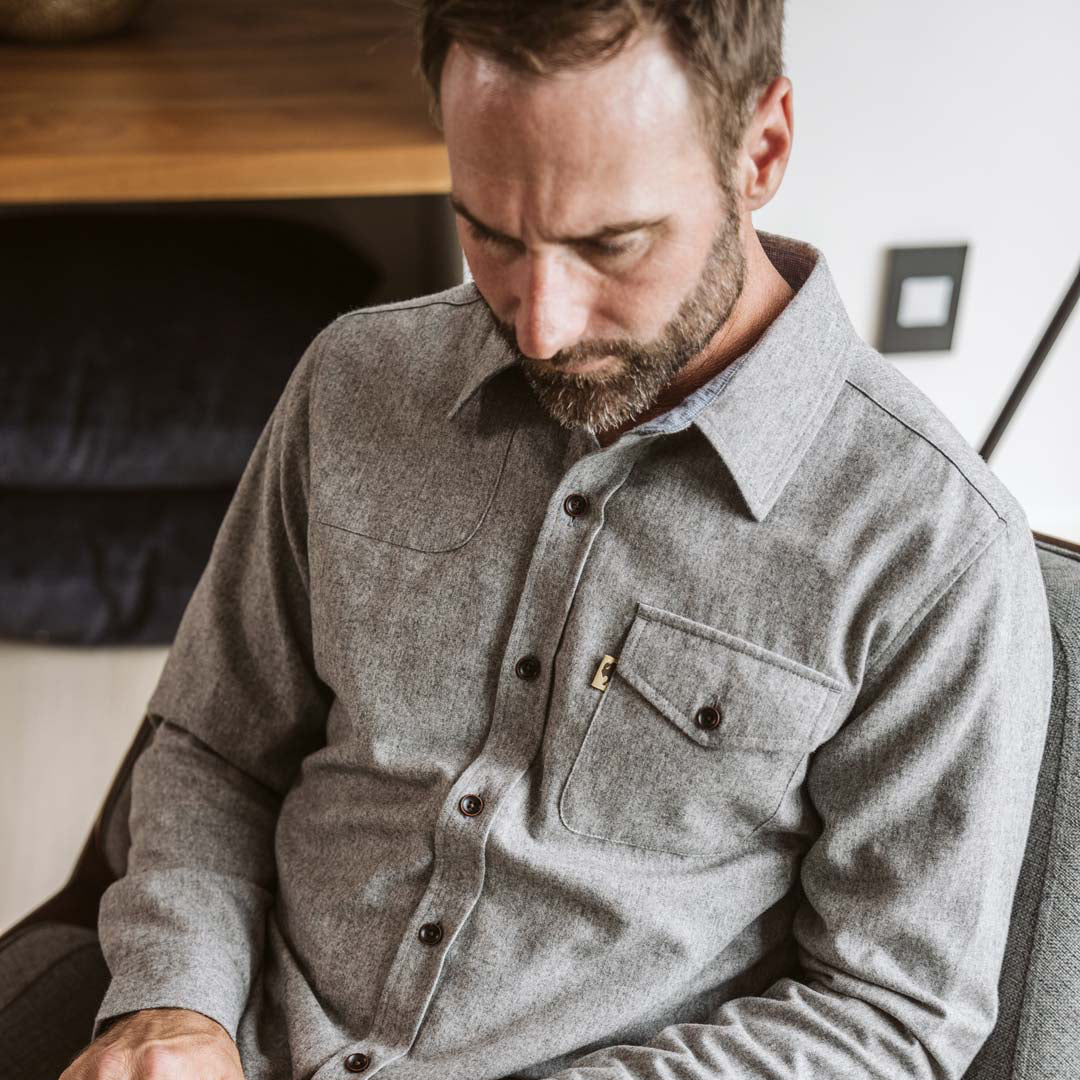 A man wearing a gray long-sleeve button-up shirt sits in a chair and types on a laptop in a modern indoor setting.