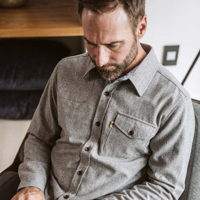 A man wearing a gray long-sleeve button-up shirt sits in a chair and types on a laptop in a modern indoor setting.