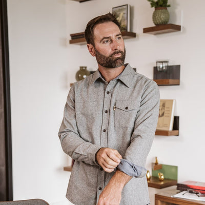 A man wearing a light gray long-sleeve button-down shirt and dark pants, standing in a modern living room setting.