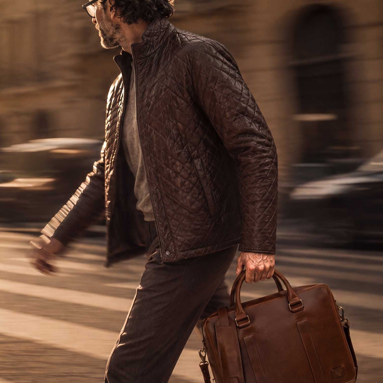 A man walking across a city crosswalk at golden hour, wearing a quilted brown jacket and carrying a brown leather briefcase.