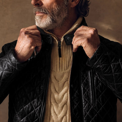Man fastening the collar of the Highlands Quilted Leather Jacket in Black over a cream cable-knit sweater.