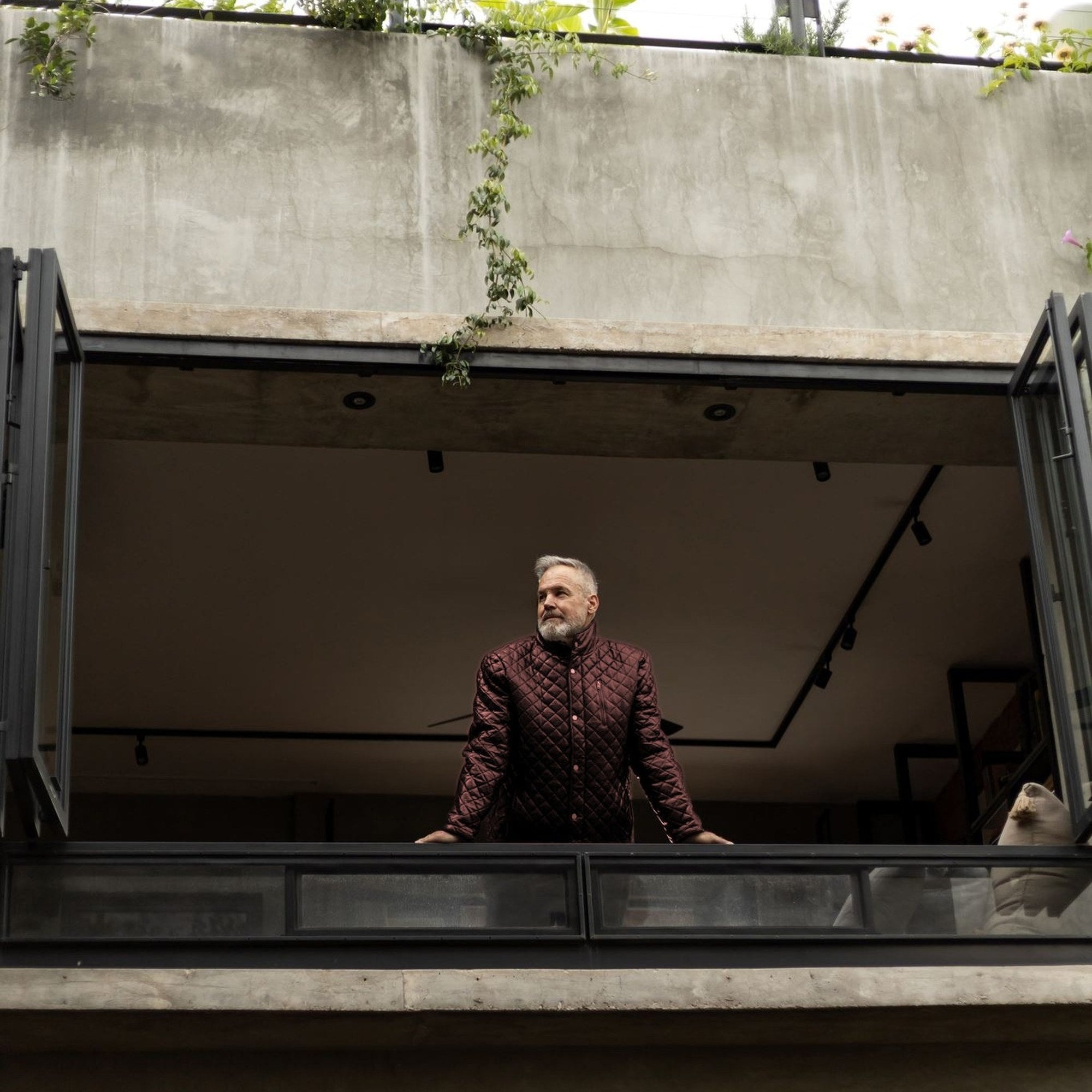 Man in the Highlands Quilted Leather Jacket in Mahogany Brown on an open-air balcony.