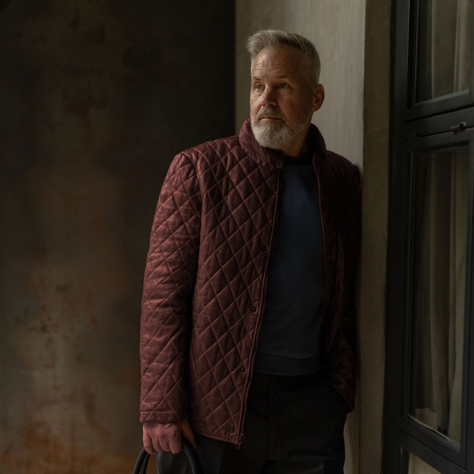 Man in the Highlands Quilted Leather Jacket in Mahogany Brown by a window, carrying a black leather briefcase.