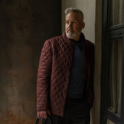 Man in the Highlands Quilted Leather Jacket in Mahogany Brown by a window, carrying a black leather briefcase.