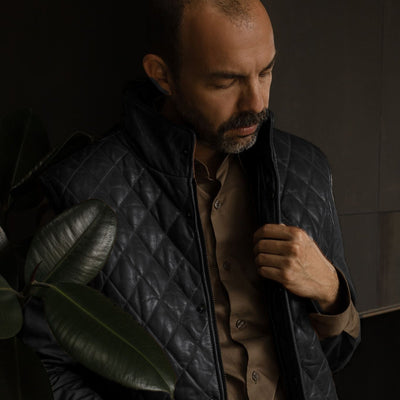 Man in the Highlands Quilted Leather Vest in Black in a dark room with plants.