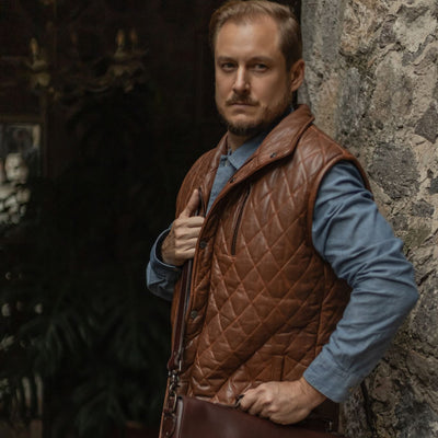 Man in the Highlands Quilted Leather Vest in Whiskey Brown under a stone archway, carrying a leather briefcase.