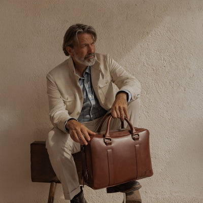 Man in a cream linen jacket seated against a sunlit wall, holding the Jefferson Leather Briefcase in Elderwood Brown.