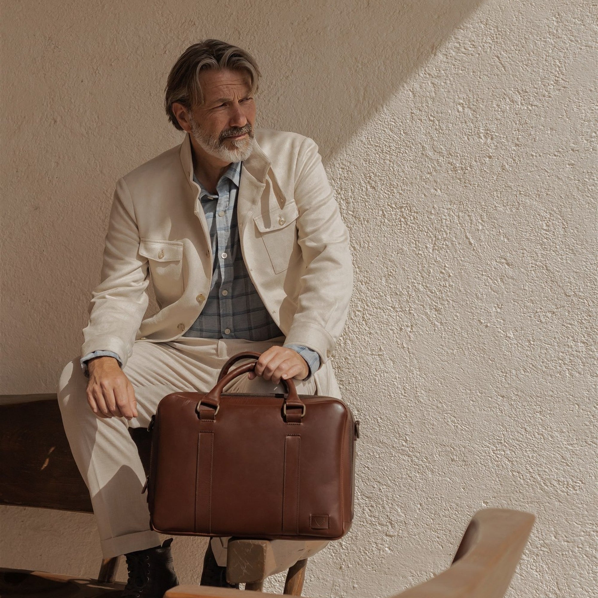 Man in a cream linen jacket seated with the Jefferson Leather Briefcase in Elderwood Brown.