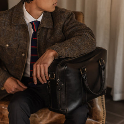 Man in a tweed jacket seated in a leather armchair with the Jefferson Leather Briefcase in Vintage Black.