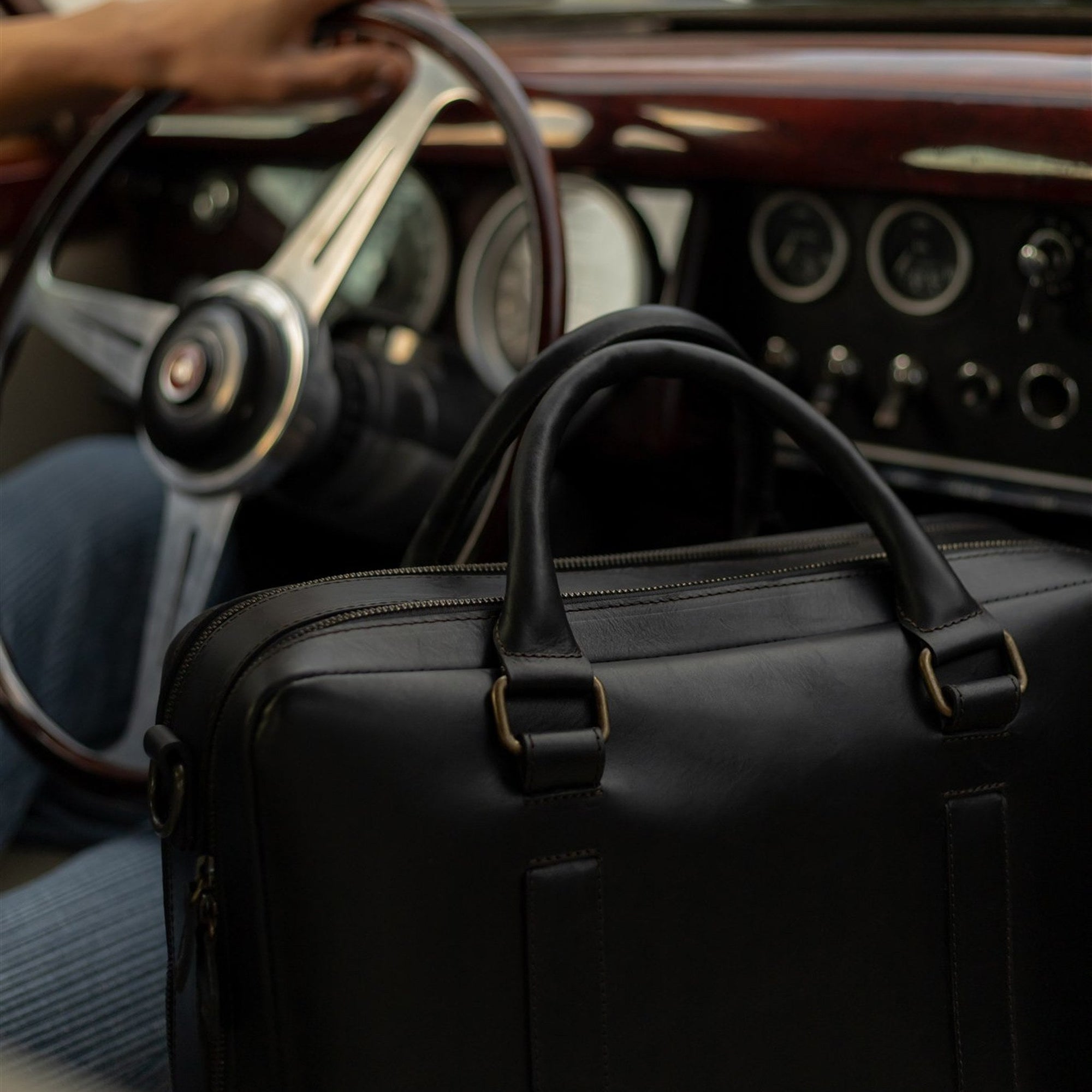 The Jefferson Leather Briefcase in Vintage Black on the front seat of a vintage car beside the driver.