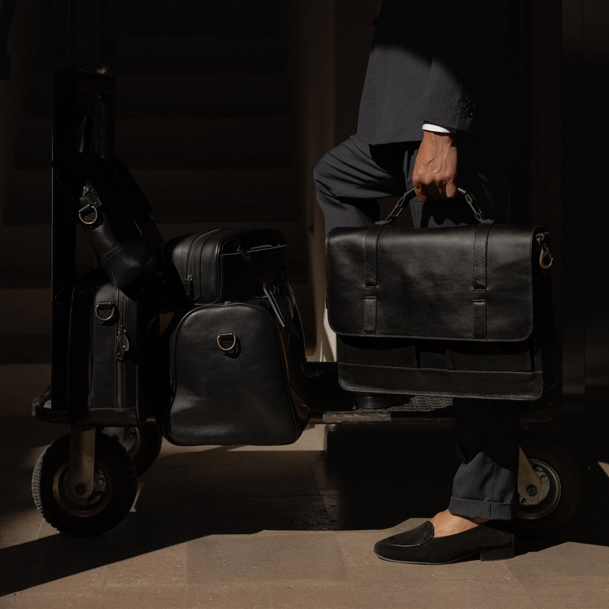 Man holding a Jefferson leather bag beside a hotel luggage cart loaded with the Jefferson collection in Vintage Black.