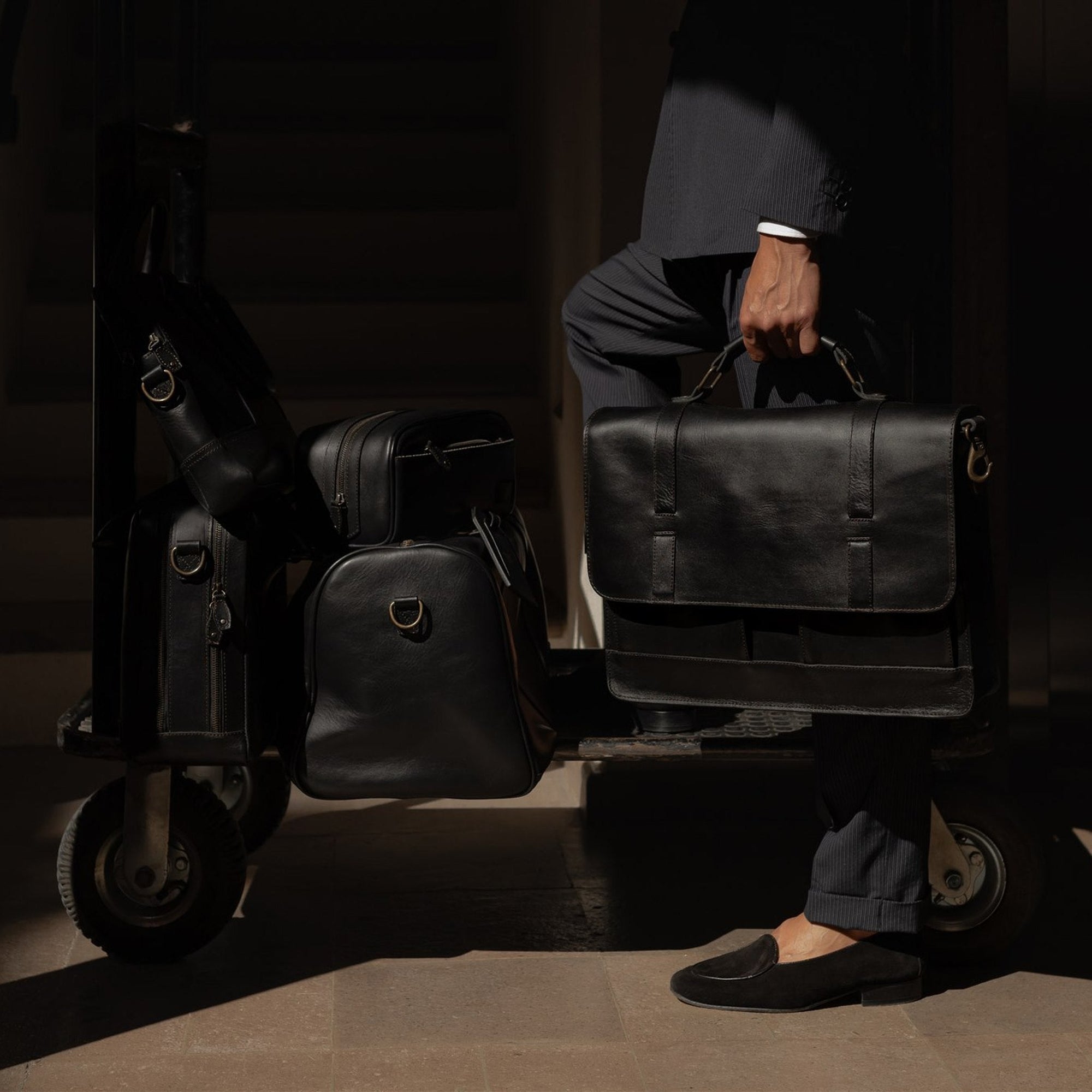 Man holding a Jefferson leather bag beside a hotel luggage cart loaded with the Jefferson collection in Vintage Black.