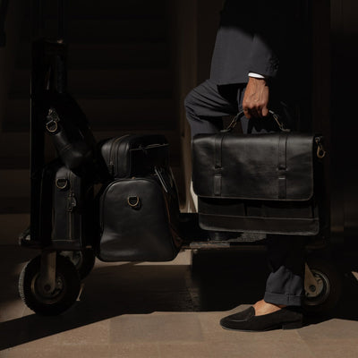 Man holding a Jefferson leather bag beside a hotel luggage cart loaded with the Jefferson collection in Vintage Black.