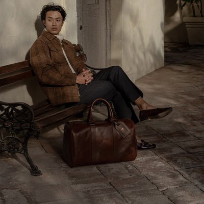 Man in a brown wool jacket seated on a bench beside the Jefferson Leather Duffle in Elderwood Brown