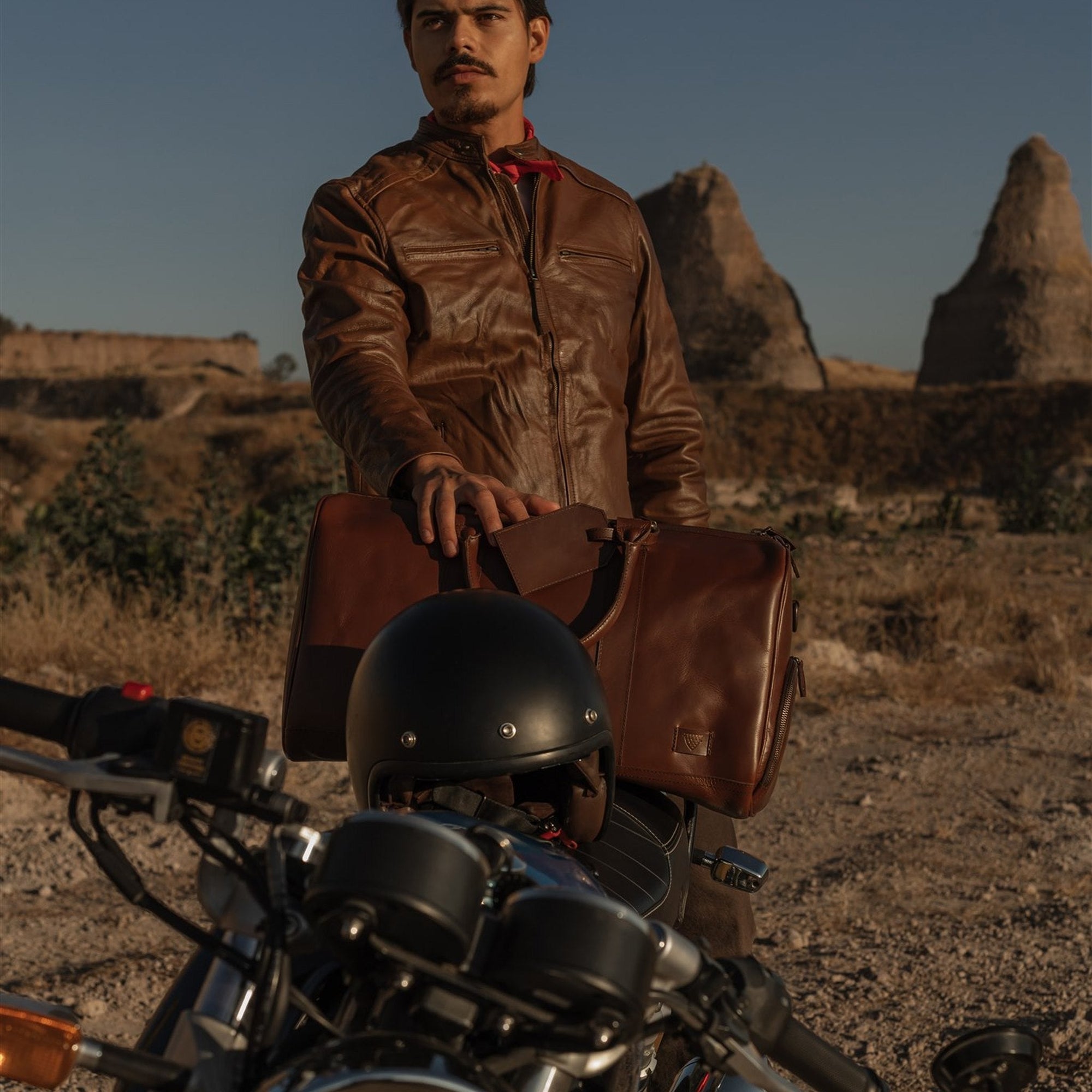 Man resting a hand on the Jefferson Leather Duffle in Elderwood Brown behind a motorcycle in the desert.