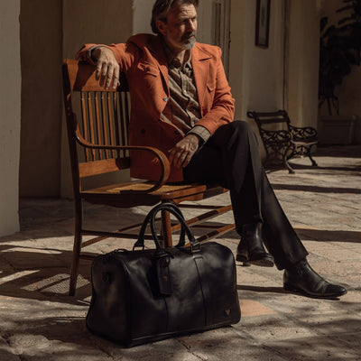Man seated on a bench in an arcade with the Jefferson Leather Duffle in Vintage Black at his feet.