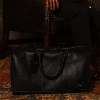 Jefferson Leather Duffle in Vintage Black on a patterned rug beside a leather chair.