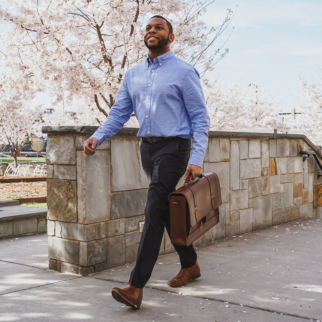 Man in a blue shirt and dark pants holding a brown briefcase, standing against a stone wall with cherry blossom trees in the background.