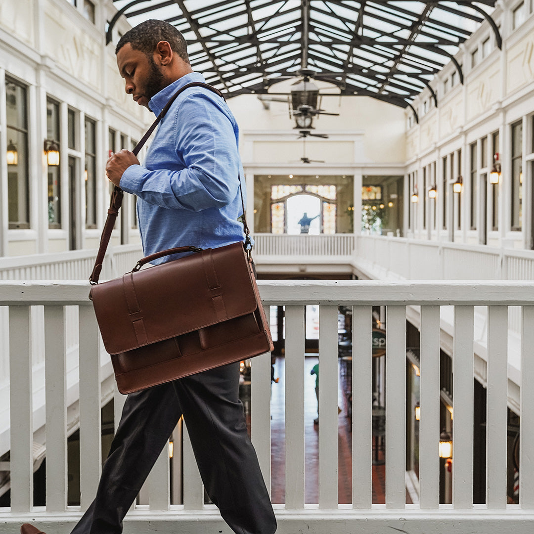 Man carrying a brown leather briefcase in an elegant indoor setting with high ceilings and large windows.