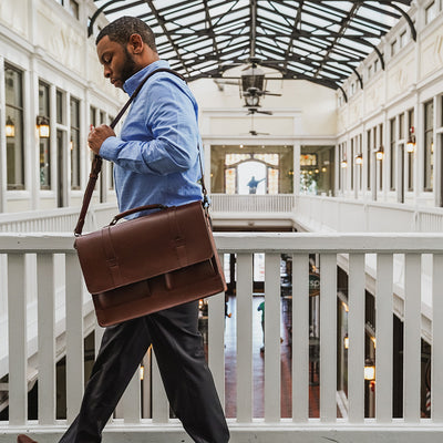 Man carrying a brown leather briefcase in an elegant indoor setting with high ceilings and large windows.