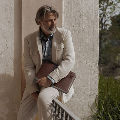 Man in a cream linen jacket seated against a wall with the Jefferson Leather Travel Portfolio in Elderwood Brown.