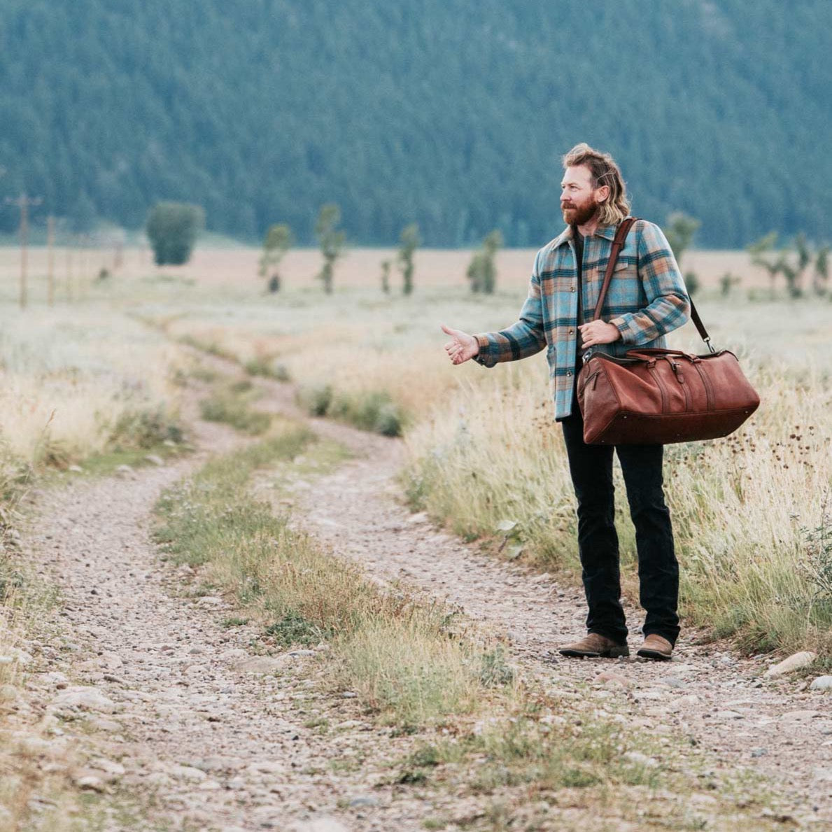Vintage brown leather duffle bag held by a traveler in a scenic Jackson Hole meadow, highlighting the rugged, stylish design.